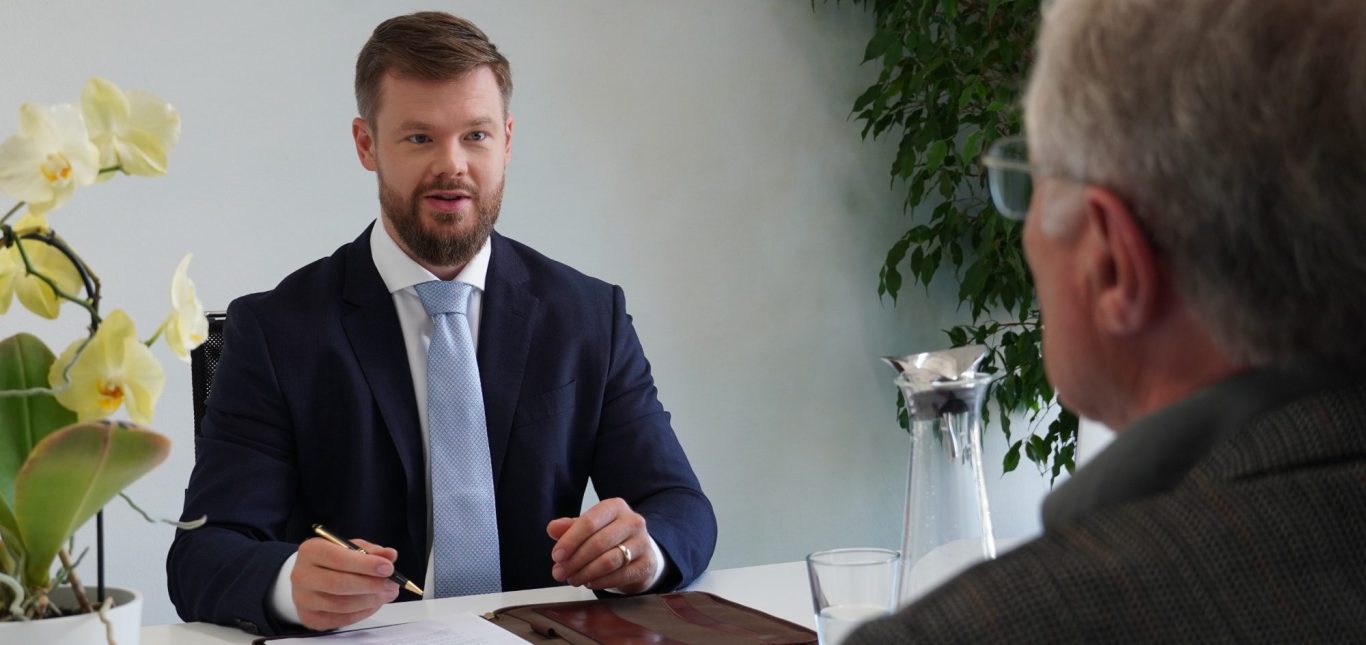 Ein Anwalt in Anzug sitzt am Tisch und spricht mit einem Mandanten. Pflanzen im Hintergrund.