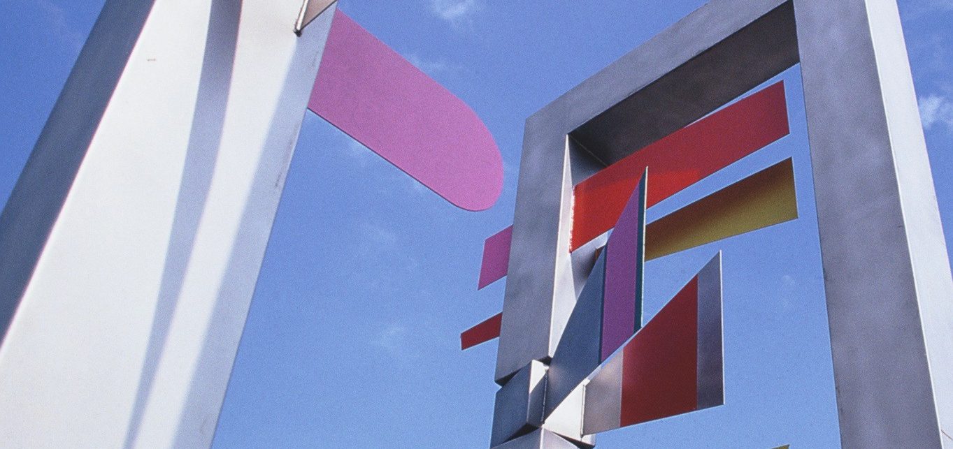 Moderne Skulptur aus Metall mit bunten, in verschiedene Richtungen zeigenden Wegweisern vor blauem Himmel.